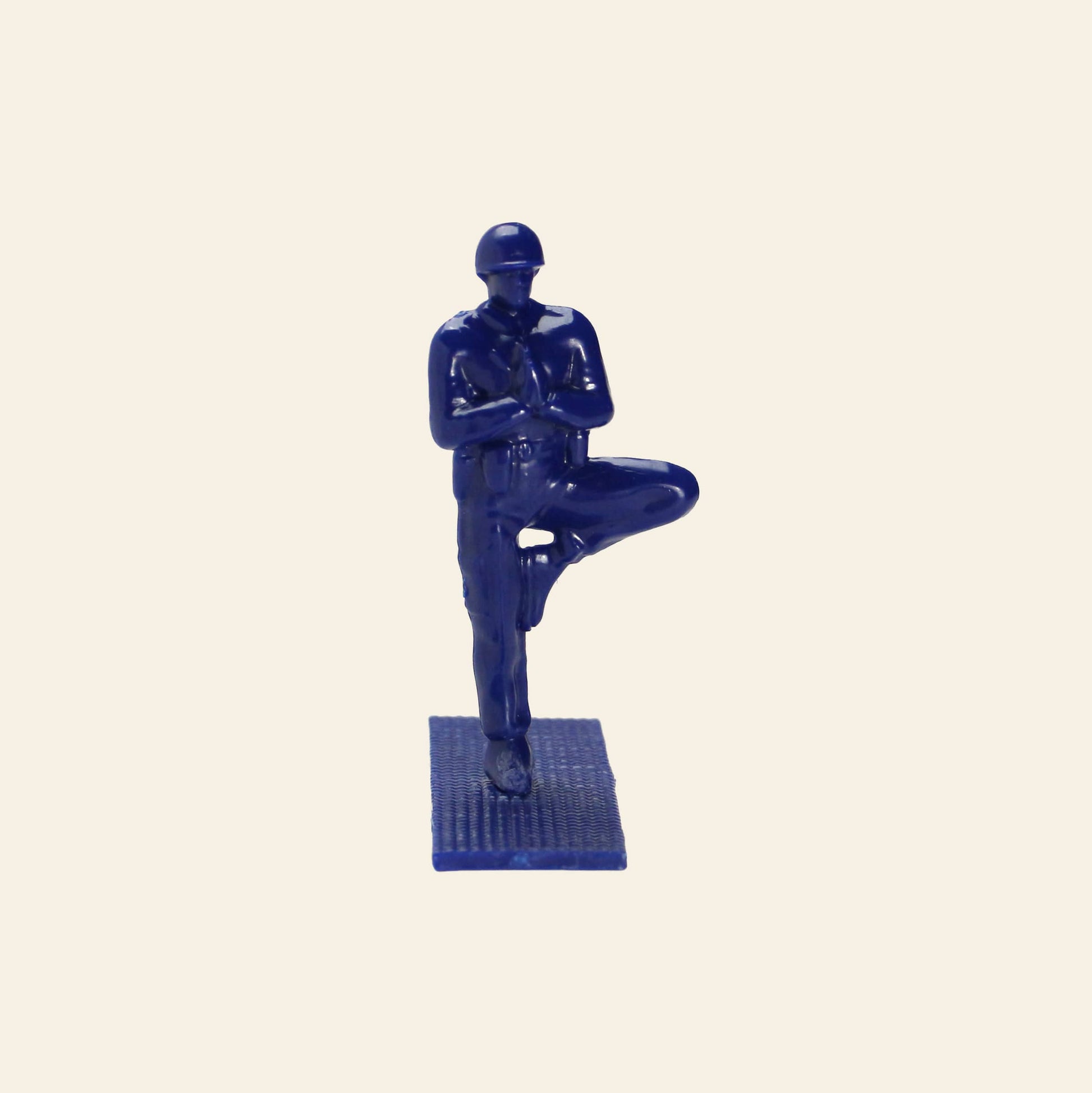 A blue plastic DETAIL Yoga Joes figure by DETAIL stands on a textured base, balancing on one leg with arms folded across the chest, capturing the playful spirit of toy soldiers doing yoga against a plain beige background.