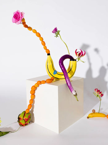 A surreal home decor scene: the DOIY Banana Vase by DOIY, bananas, orange kumquats, pink roses, and other flowers are artistically arranged on a white cube against a white background.