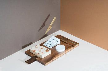 The DOIY Cheeseporn cheeseboard by DOIY, crafted from acacia wood, presents three cheese blocks, a slicer, and a floating golden knife with shadow highlights against a brown and beige backdrop.