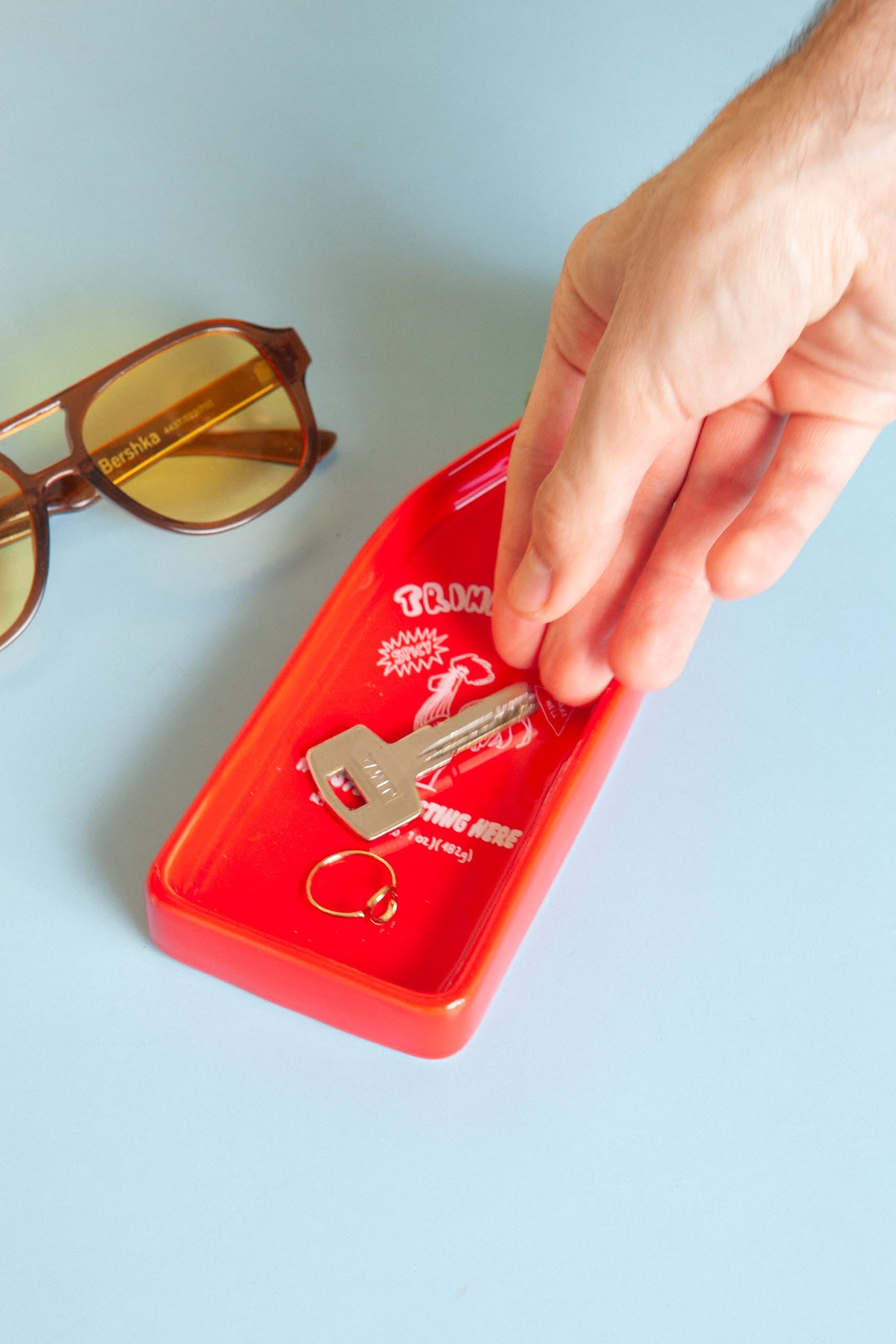 A hand reaches for a key in the DOIY Corner Shop Sriracha Trinket Tray by DOIY, which also holds a key ring and sits next to brown sunglasses on a light blue surface.