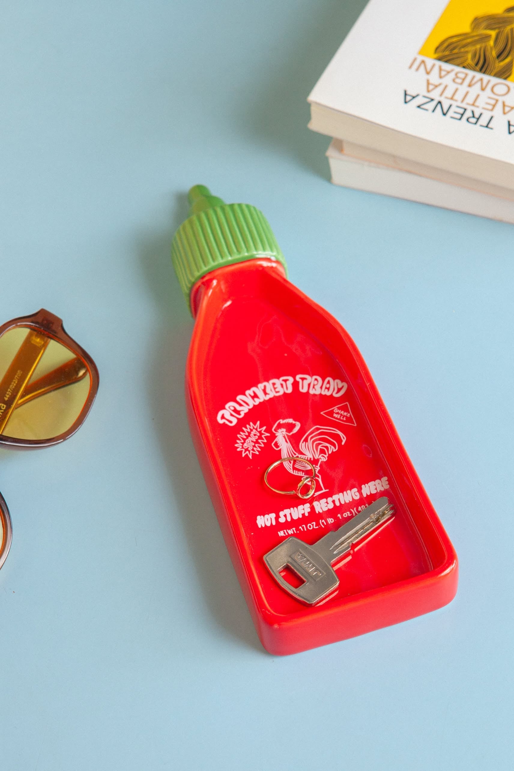 The DOIY Corner Shop Sriracha Trinket Tray by DOIY, shaped like the iconic bottle with green cap, features TRINKET TRAY, HOT STUFF RESTING HERE, and a rooster graphic—perfect for holding keys beside sunglasses and books.