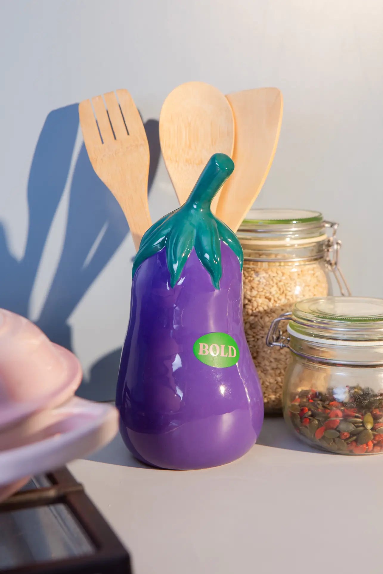 The DOIY Farmers’ Market Eggplant Utensils Pot by DOIY sits on the kitchen counter, bringing playful vegetable-themed decor to your space among wooden utensils and jars of grains and spices.