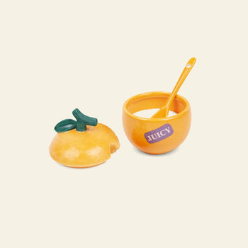 The DOIY Farmers’ Market Orange Sugar Bowl by DOIY features a round, orange ceramic design with a green stem lid, JUICY label, and yellow spoon—perfect for sauces or dips—set against a light background for a farmers market feel.