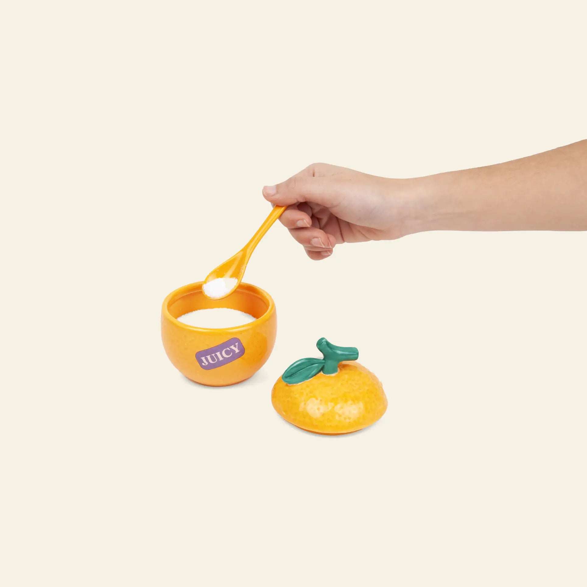 A hand holds a spoon of white powder above the DOIY Farmers’ Market Orange Sugar Bowl by DOIY, shaped like an orange with a “JUICY” label and matching green-leaf lid, ideal for serving sauces or dips on a light background.