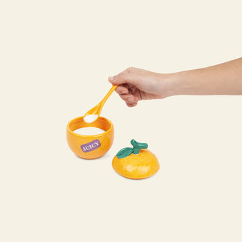 A hand holds a spoon of white powder above the DOIY Farmers’ Market Orange Sugar Bowl by DOIY, shaped like an orange with a “JUICY” label and matching green-leaf lid, ideal for serving sauces or dips on a light background.