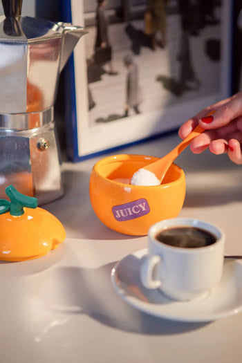 A hand with red nail polish adds sugar from the DOIY Farmers’ Market Orange Sugar Bowl—a ceramic piece by DOIY with a leafy lid—into coffee, while a moka pot and the bowl’s orange leaf-shaped lid rest on the table.