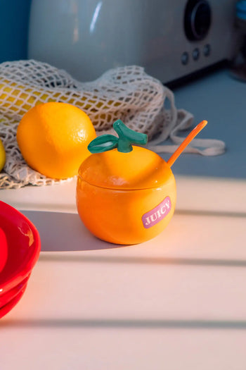 The DOIY Farmers’ Market Orange Sugar Bowl by DOIY features a round orange ceramic design with a green leaf-shaped lid and matching spoon. A “JUICY” sticker decorates the bowl, while sunlight casts playful shadows over lemon and mesh bag in the background.