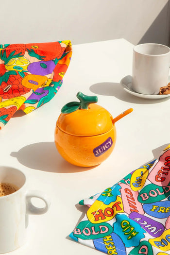 The DOIY Farmers’ Market Orange Sugar Bowl by DOIY, shaped like a bright orange and labeled “JUICY,” sits on a white table with colorful napkins and coffee cups, ideal for serving sauces or dips—evoking farmers’ market charm in the sunlight.