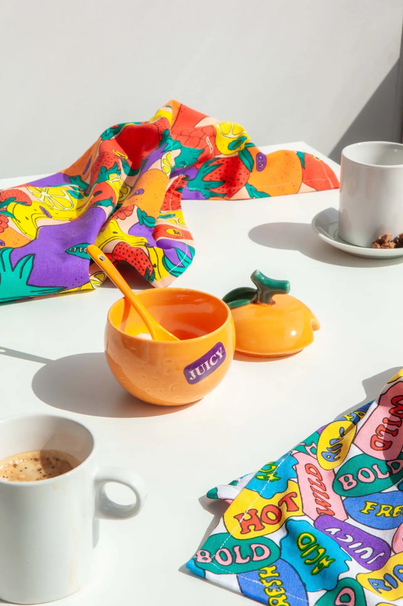 A vibrant table with fruit-themed napkins, two white coffee cups, and the DOIY Farmers’ Market Orange Sugar Bowl—ideal for sauces and dips—creates a sunny, cheerful farmers market vibe.