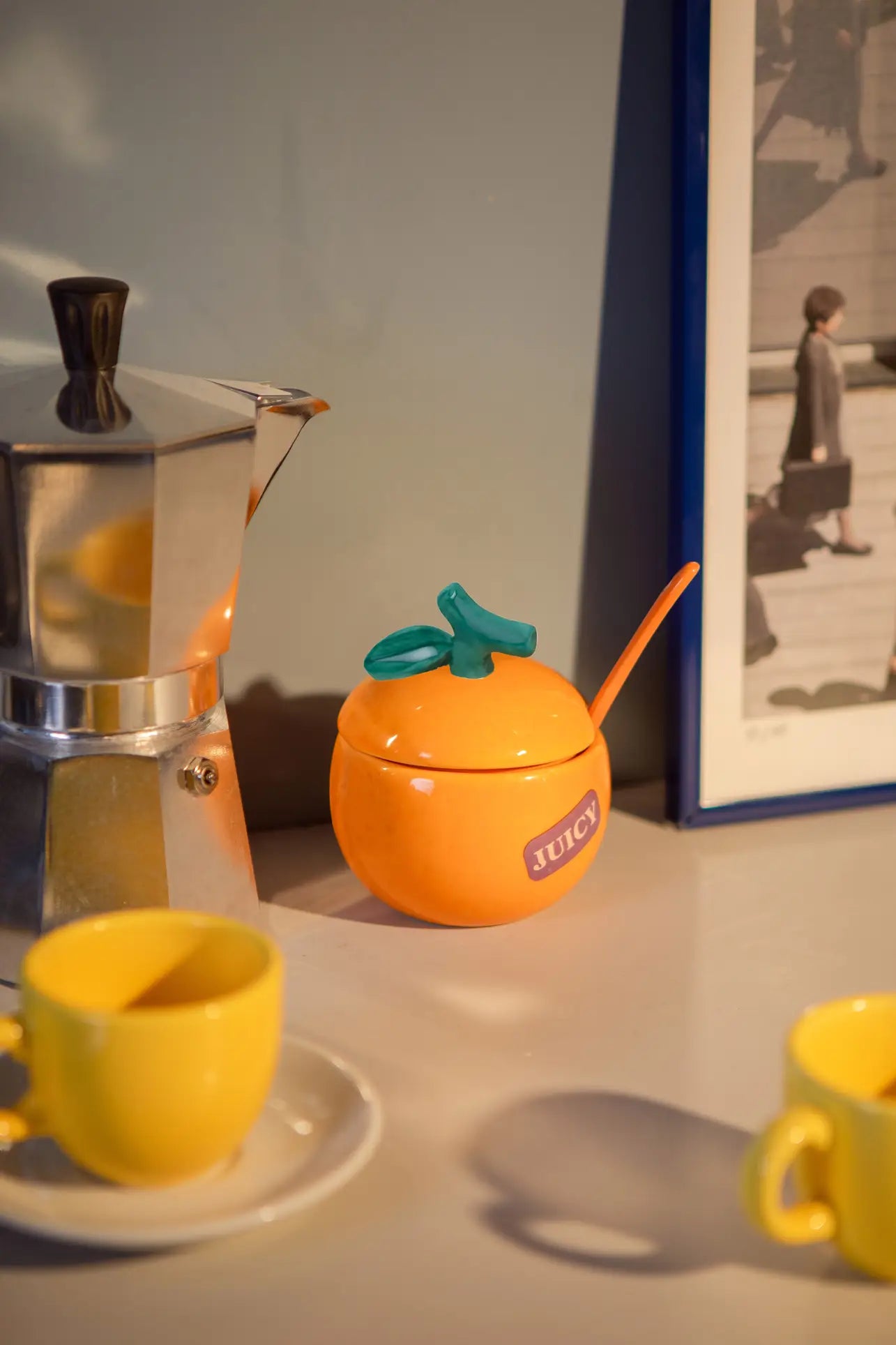 A silver moka pot, a DOIY Farmers’ Market Orange Sugar Bowl with spoon, and two yellow cups rest on a countertop beside a framed photograph.