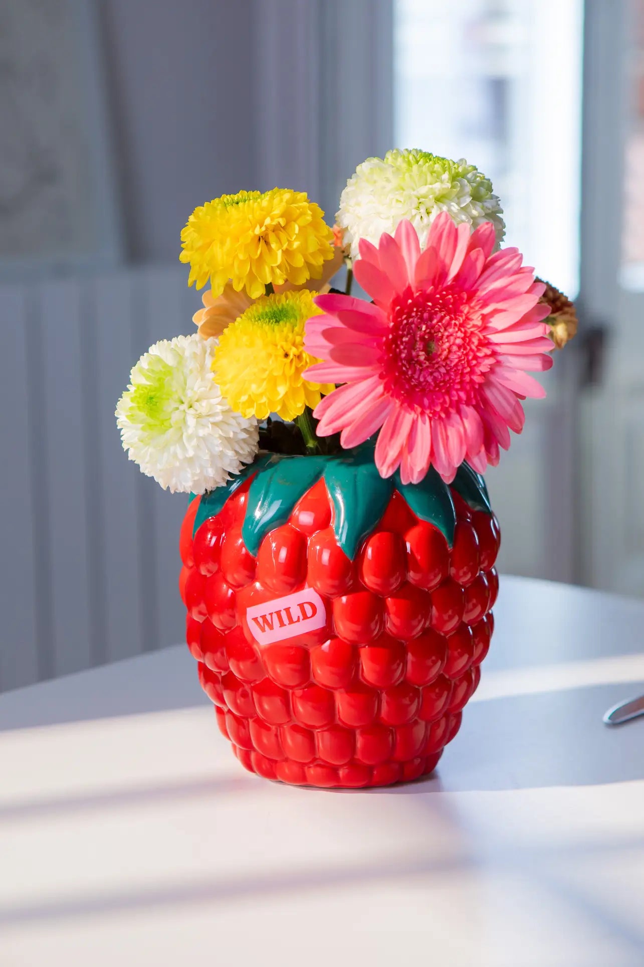The DOIY Farmers Market Raspberry Vase, shaped like a red strawberry and labeled WILD, displays pink gerbera daisies and pompom blooms, bringing playful style and vibrant charm to your home decor on a sunlit white table.