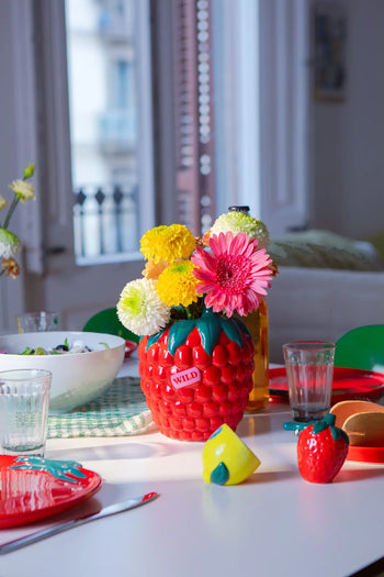 A cheerful table setting features the DOIY Farmers Market Raspberry Vase by DOIY, paired with a matching strawberry-shaped salt shaker. Sunlight streams in, highlighting the vibrant charm of this playful decor accent.