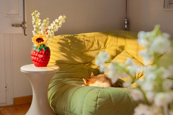 A cat naps on a light green couch in a sunlit room. A DOIY Farmers Market Raspberry Vase from DOIY, filled with sunflowers and white blooms, sits on a white side table, adding vibrant charm to the inviting decor. White blossoms complete the scene.