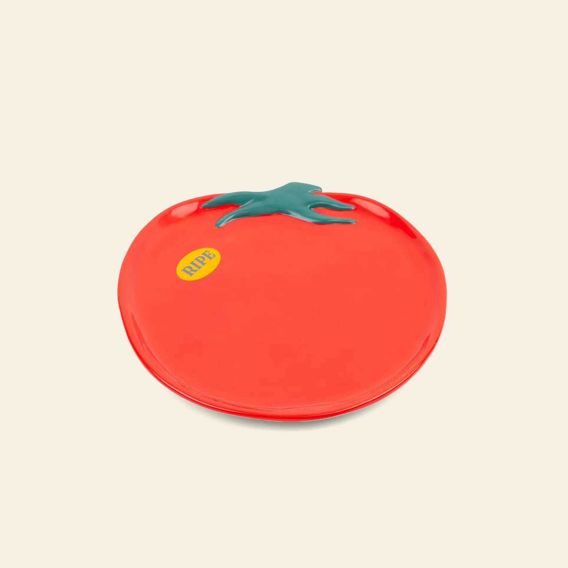 Create a unique table setting with the DOIY Farmers Market Tomato Plate by DOIY—a ceramic plate shaped like a red tomato with a green stem and yellow RIPE sticker, displayed on a light beige background.