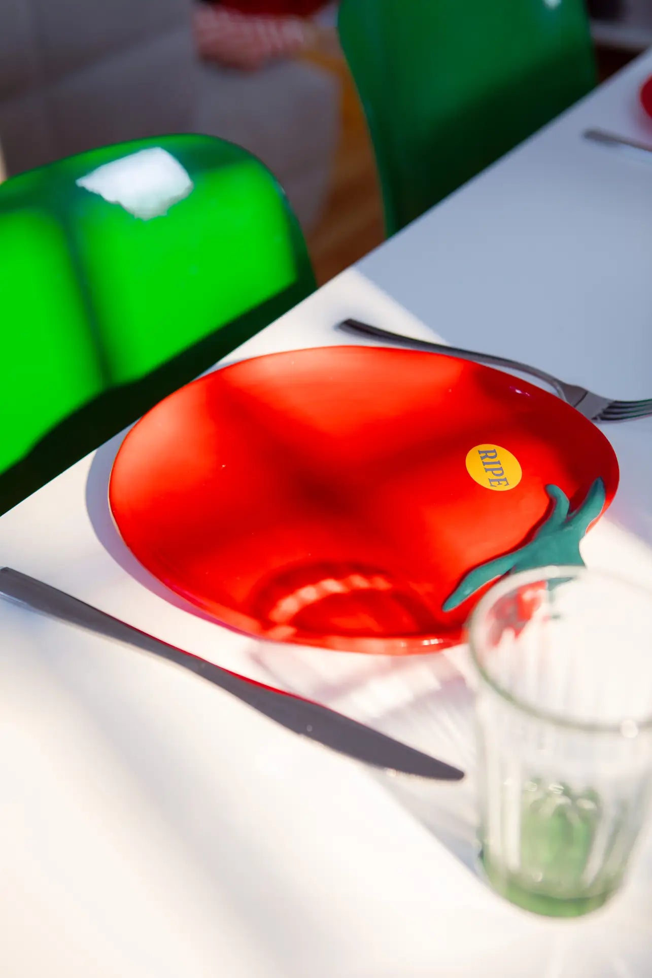 The DOIY Farmers Market Tomato Plate, a red ceramic plate shaped like a tomato with a ripe sticker by DOIY, sits on a white table with cutlery and glassware, surrounded by bright green chairs.