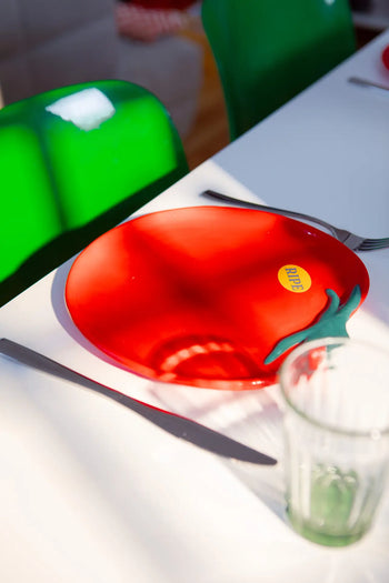 The DOIY Farmers Market Tomato Plate, a red ceramic plate shaped like a tomato with a ripe sticker by DOIY, sits on a white table with cutlery and glassware, surrounded by bright green chairs.