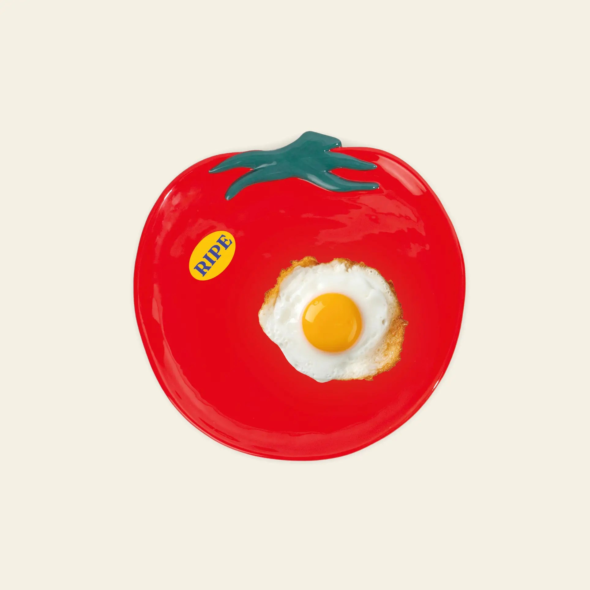 A fried egg rests on the DOIY Farmers Market Tomato Plate, a ceramic piece by DOIY shaped like a tomato with green leaves and a yellow RIPE sticker, set against an off-white background for a charming table setting.