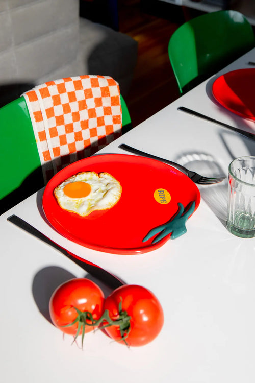 A unique table setting showcases the DOIY Farmers Market Tomato Plate by DOIY, holding a sunny-side-up egg on a white table. Nearby are fresh tomatoes, a fork, glass, and a green chair draped with an orange-checkered towel.