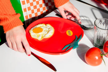 A person in an orange sweater holds cutlery near the DOIY Farmers Market Tomato Plate by DOIY, featuring a fried egg and blue chili pepper design. More tomatoes and a glass of water complete the unique table setting on the white surface.