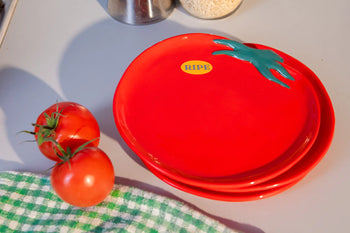 Two DOIY Farmers Market Tomato Plates by DOIY, featuring a tomato design and RIPE stickers, sit on a countertop near fresh tomatoes and a green-and-white checkered cloth—ideal for a unique table setting.