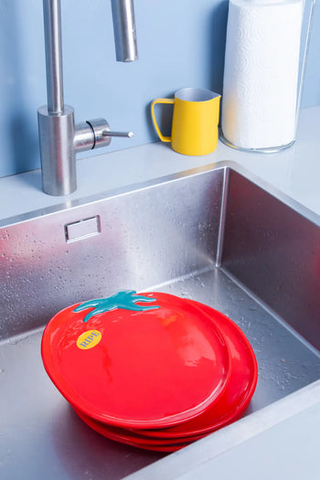 A stack of DOIY Farmers Market Tomato Plates sits in a stainless steel sink, water droplets catching the light. Nearby are a yellow mug and paper towel roll, creating a distinctive counter display.