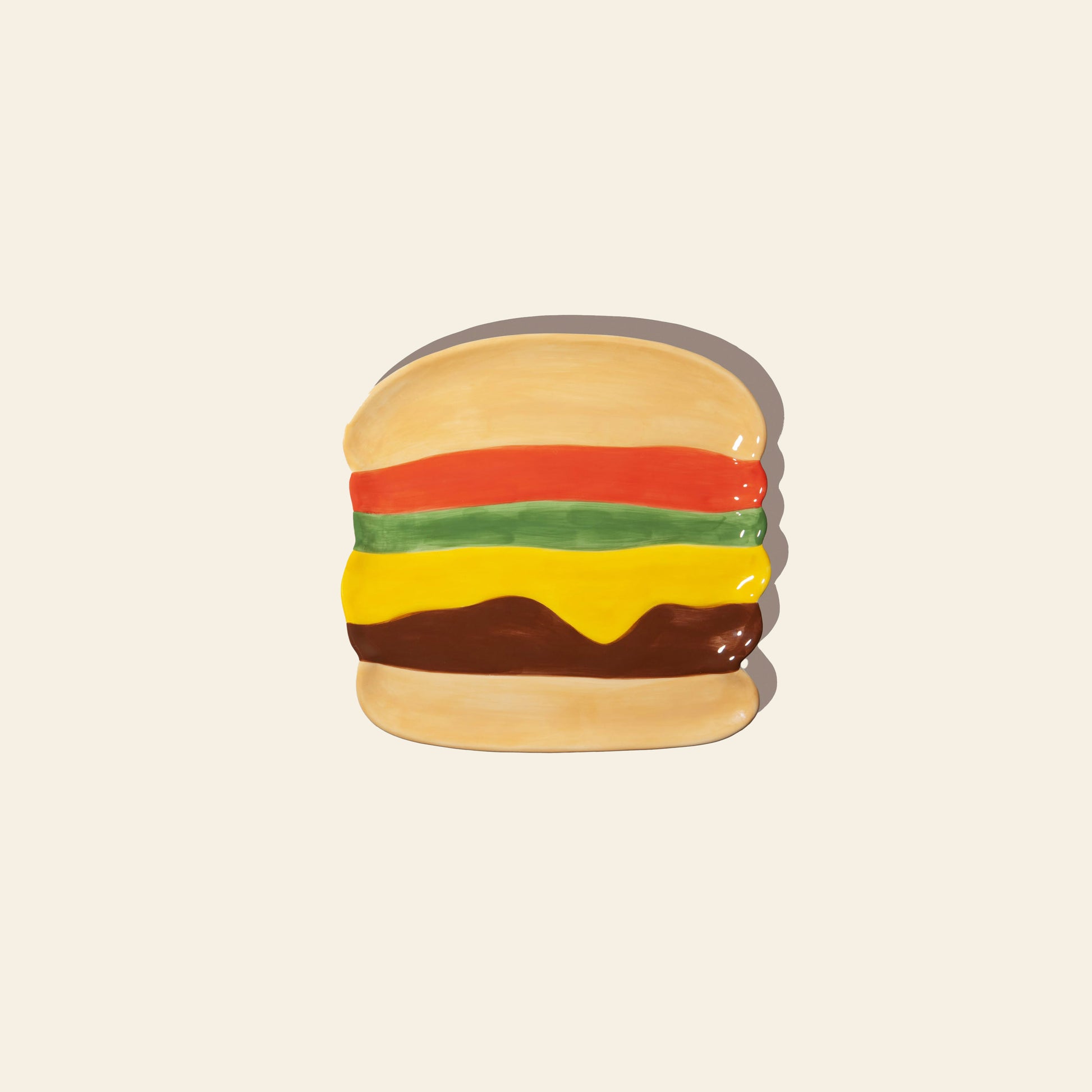 The DOIY Fast Food Burger Plate by DOIY is a ceramic plate painted to look like a cheeseburger, with layers of lettuce, tomato, cheese, and patty—adding retro diner style to your table against a plain light background.