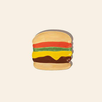 The DOIY Fast Food Burger Plate by DOIY is a ceramic plate painted to look like a cheeseburger, with layers of lettuce, tomato, cheese, and patty—adding retro diner style to your table against a plain light background.