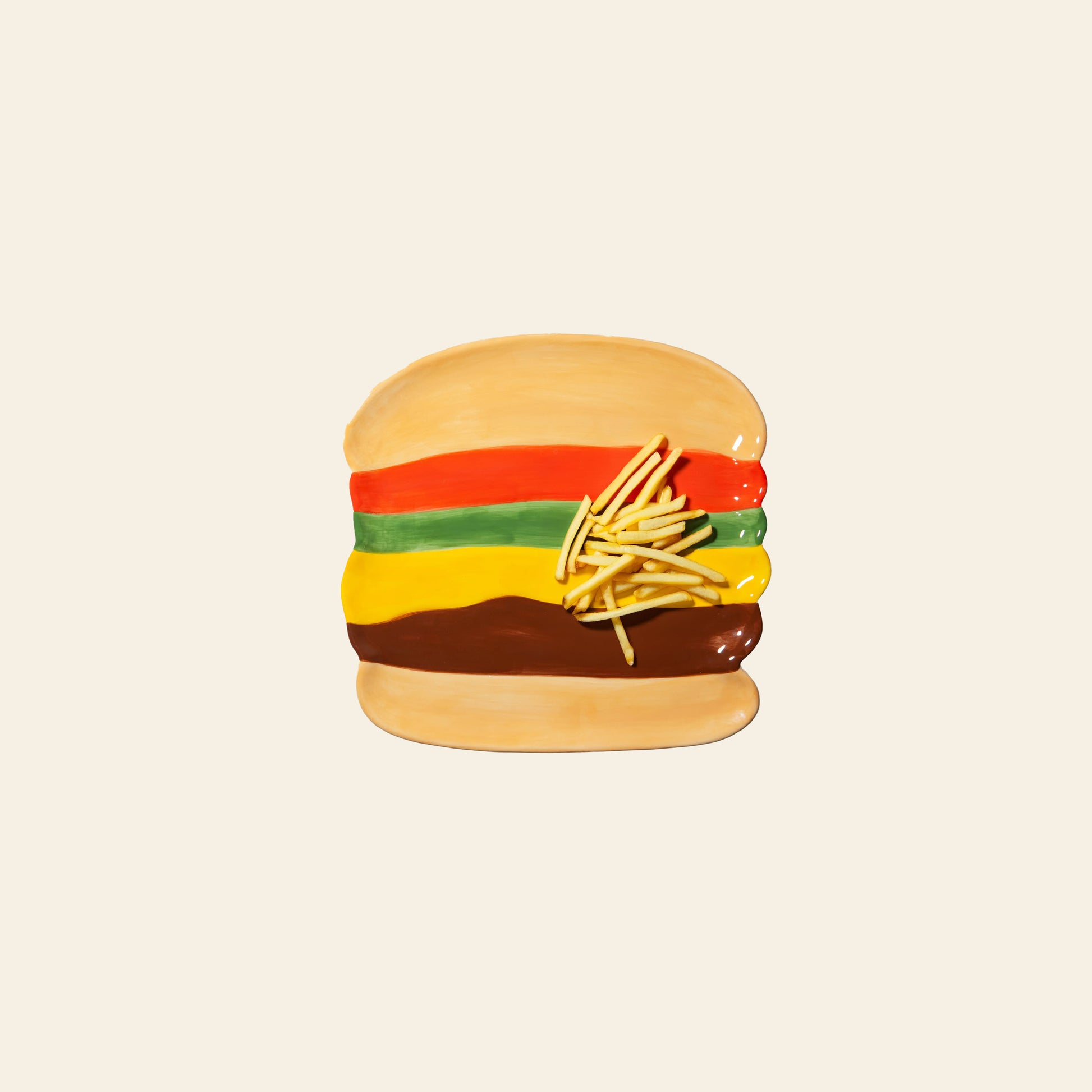The DOIY Fast Food Burger Plate by DOIY features a ceramic plate shaped like a hamburger with bun, lettuce, tomato, cheese, patty, and ceramic fries on top. Set against a plain beige background, it adds retro charm to your table.