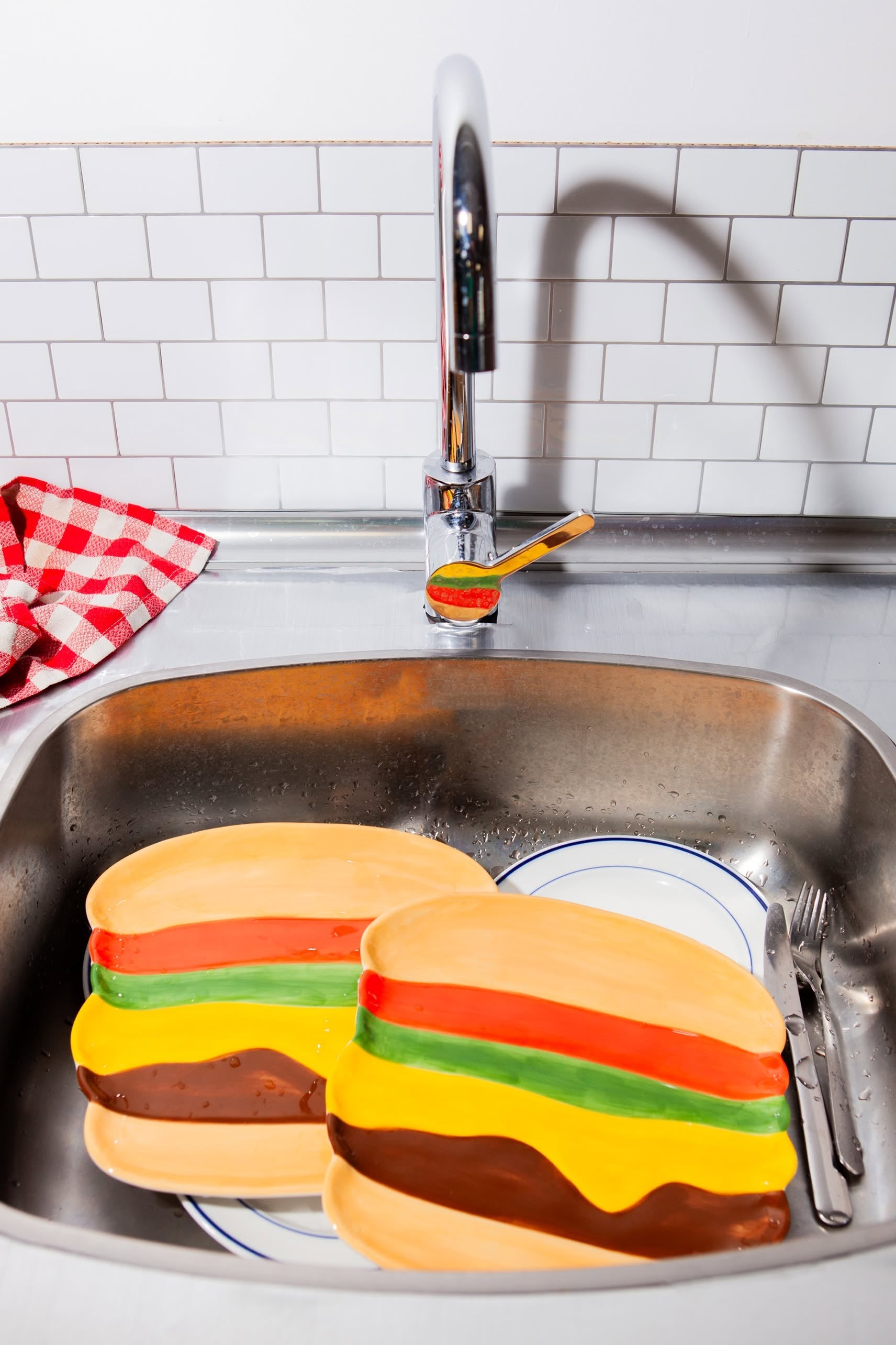 Two DOIY Fast Food Burger Plates from DOIY, featuring colorful retro designs, sit by the kitchen sink with cutlery and a red-and-white checkered towel. Nearby, a toothbrush with toothpaste adds to the vintage diner atmosphere.