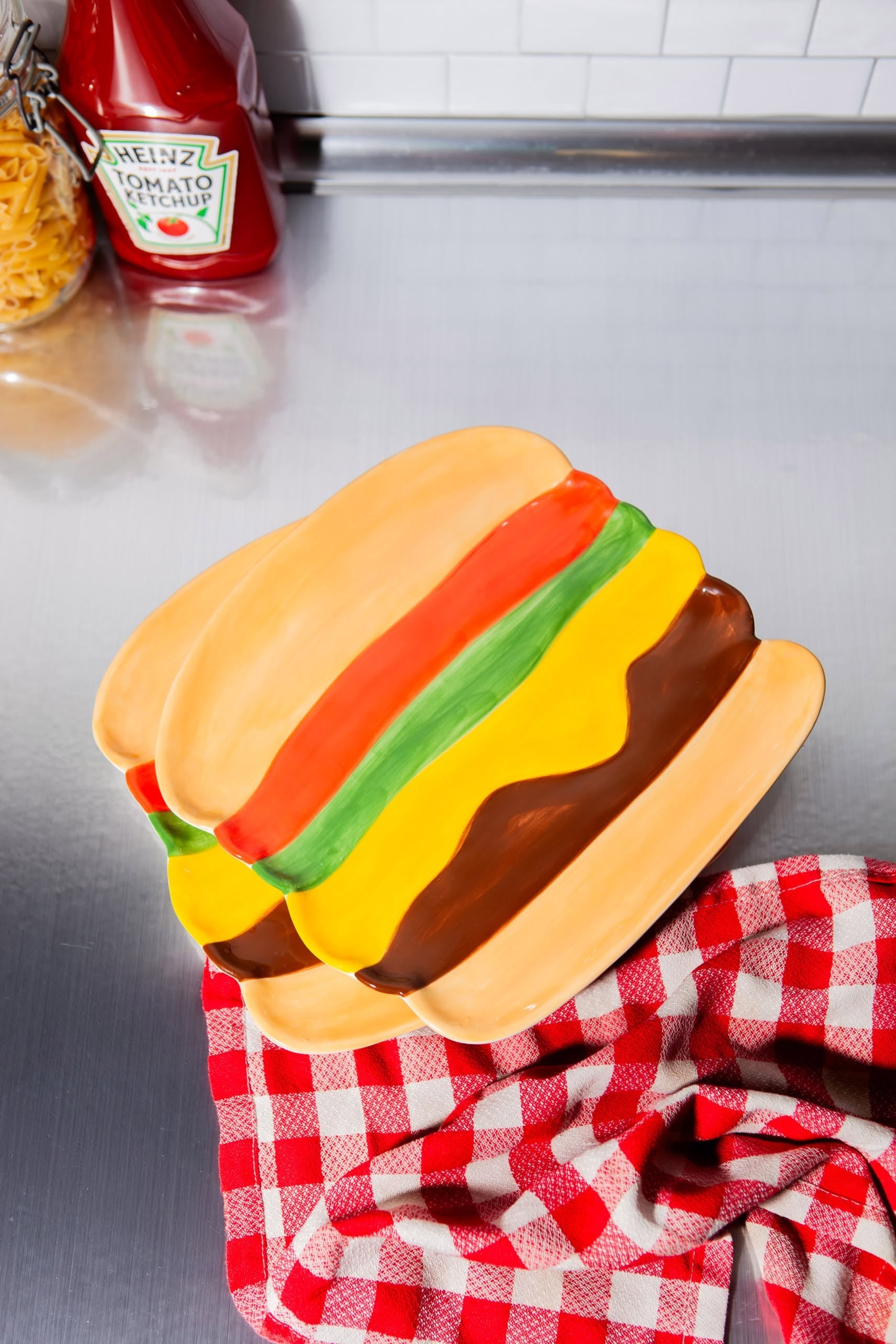 The DOIY Fast Food Burger Plate by DOIY boasts retro flair with colorful sections inspired by burger ingredients, displayed on a red checkered cloth atop a metal counter, with a ketchup bottle and pasta jar in the background.
