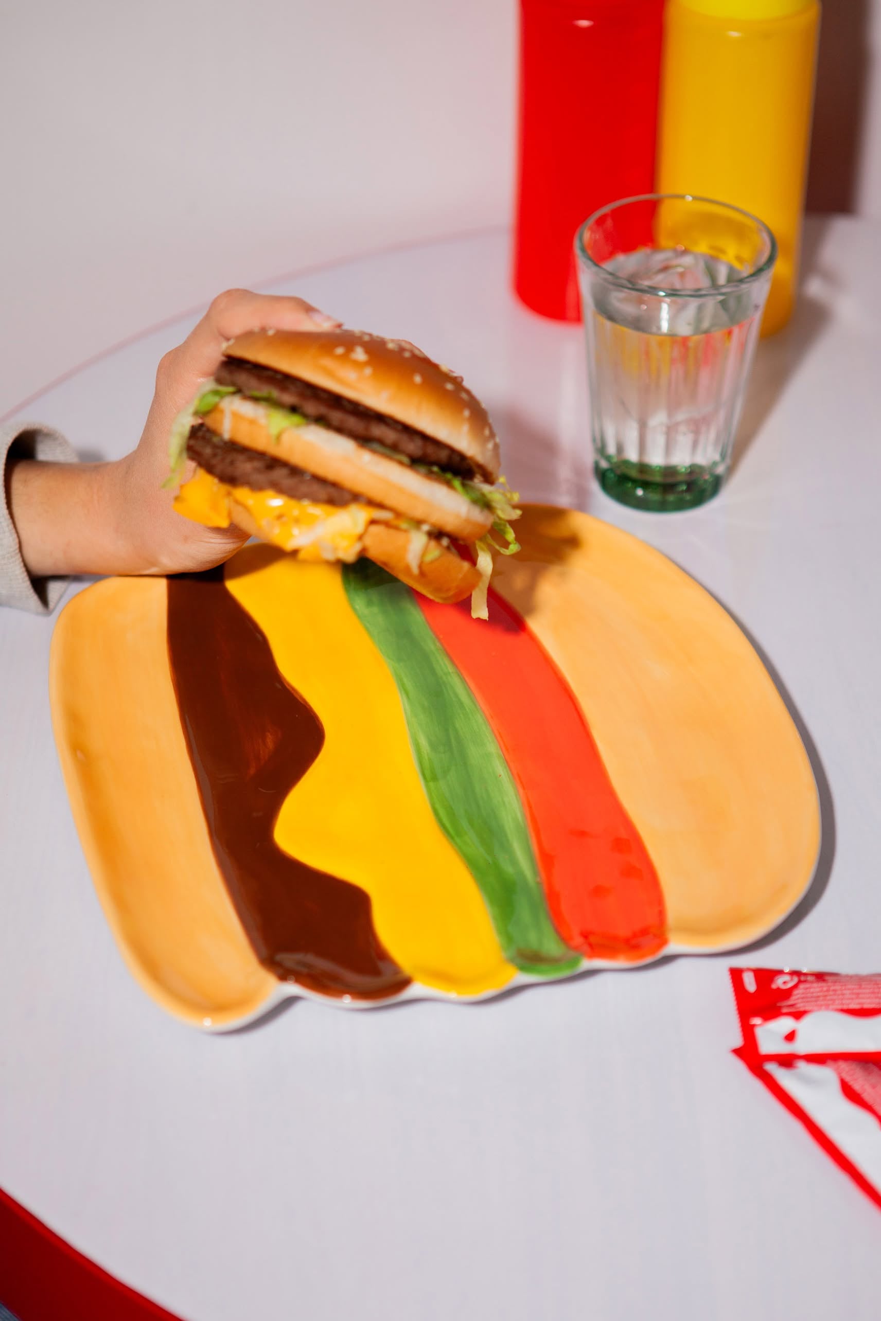 A hand holds a cheeseburger above the DOIY Fast Food Burger Plate by DOIY, featuring colorful stripes inspired by burger toppings. On the table are a glass of water, ketchup and mustard bottles, and a torn ketchup packet.