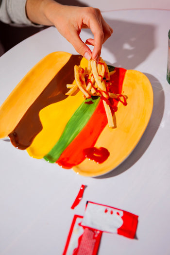A hand picks up French fries from the DOIY Fast Food Burger Plate by DOIY, featuring colorful striped sauces evoking retro charm. Two opened ketchup packets rest on the nearby white table.