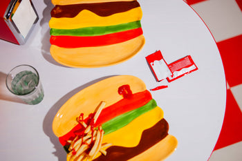 A retro white table features two DOIY Fast Food Burger Plates by DOIY—cheeseburger designs, one with fries and ketchup. A torn ketchup packet, water glass, and red napkin holder complete the vintage diner vibe.