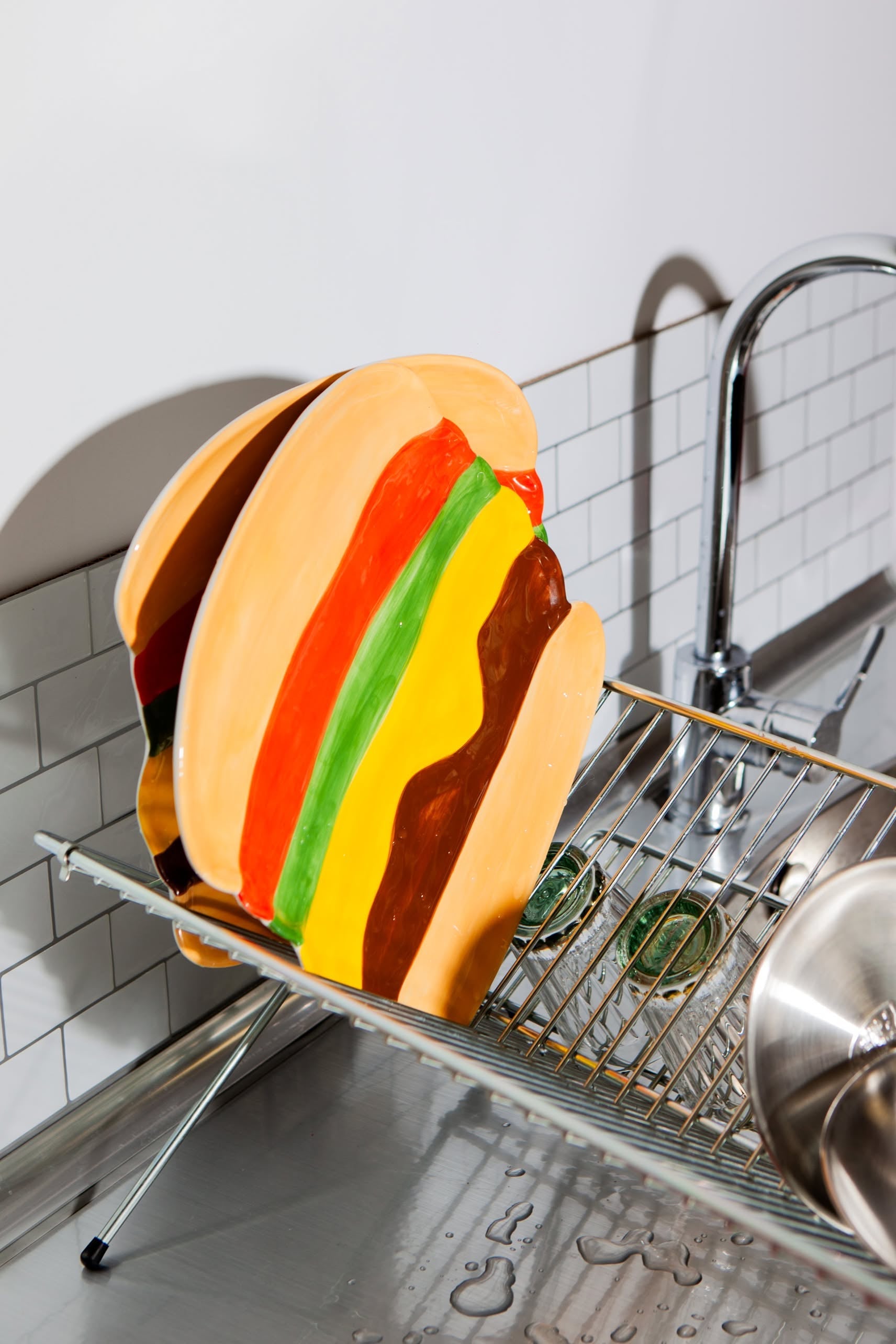 The DOIY Fast Food Burger Plate by DOIY, with its colorful retro charm, dries on a dish rack beside the kitchen sink, surrounded by glasses and water droplets shimmering on the metal surface.