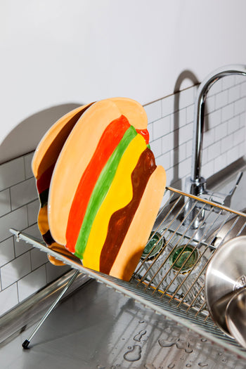 The DOIY Fast Food Burger Plate by DOIY, with its colorful retro charm, dries on a dish rack beside the kitchen sink, surrounded by glasses and water droplets shimmering on the metal surface.