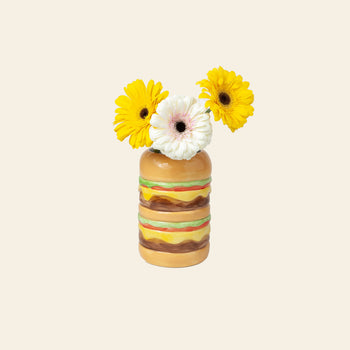 The DOIY Fast Food Burger Vase by DOIY holds three gerbera daisies—two yellow and one white with pink edges—set against a plain, light beige background.