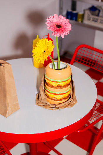 The DOIY Fast Food Burger Vase by DOIY holds two vibrant gerbera daisies on a round white table with red legs; nearby are a brown paper bag, yellow and red squeeze bottles, and red chairs.