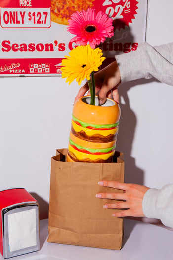 A hand places the DOIY Fast Food Burger Vase, designed like stacked burgers and filled with bright pink and yellow flowers, into a brown paper bag. A retro napkin dispenser and pizza ad enhance the scene in the background.
