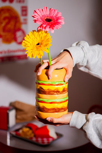 Someone holds the DOIY Fast Food Burger Vase by DOIY, designed like a stacked hamburger and filled with a yellow and pink flower. Its retro style pops against the blurred diner setting with vibrant red décor in the background.