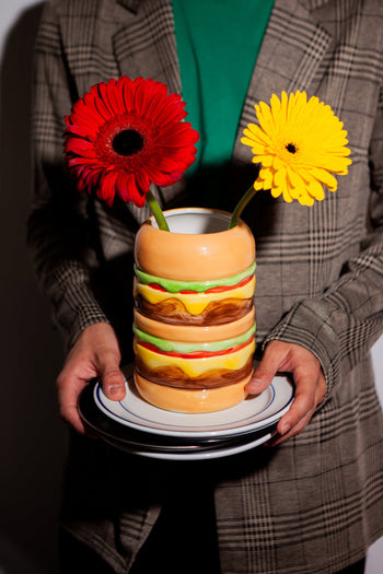 A person in a plaid blazer holds a DOIY Fast Food Burger Vase by DOIY, shaped like a burger and filled with red and yellow gerbera daisies.