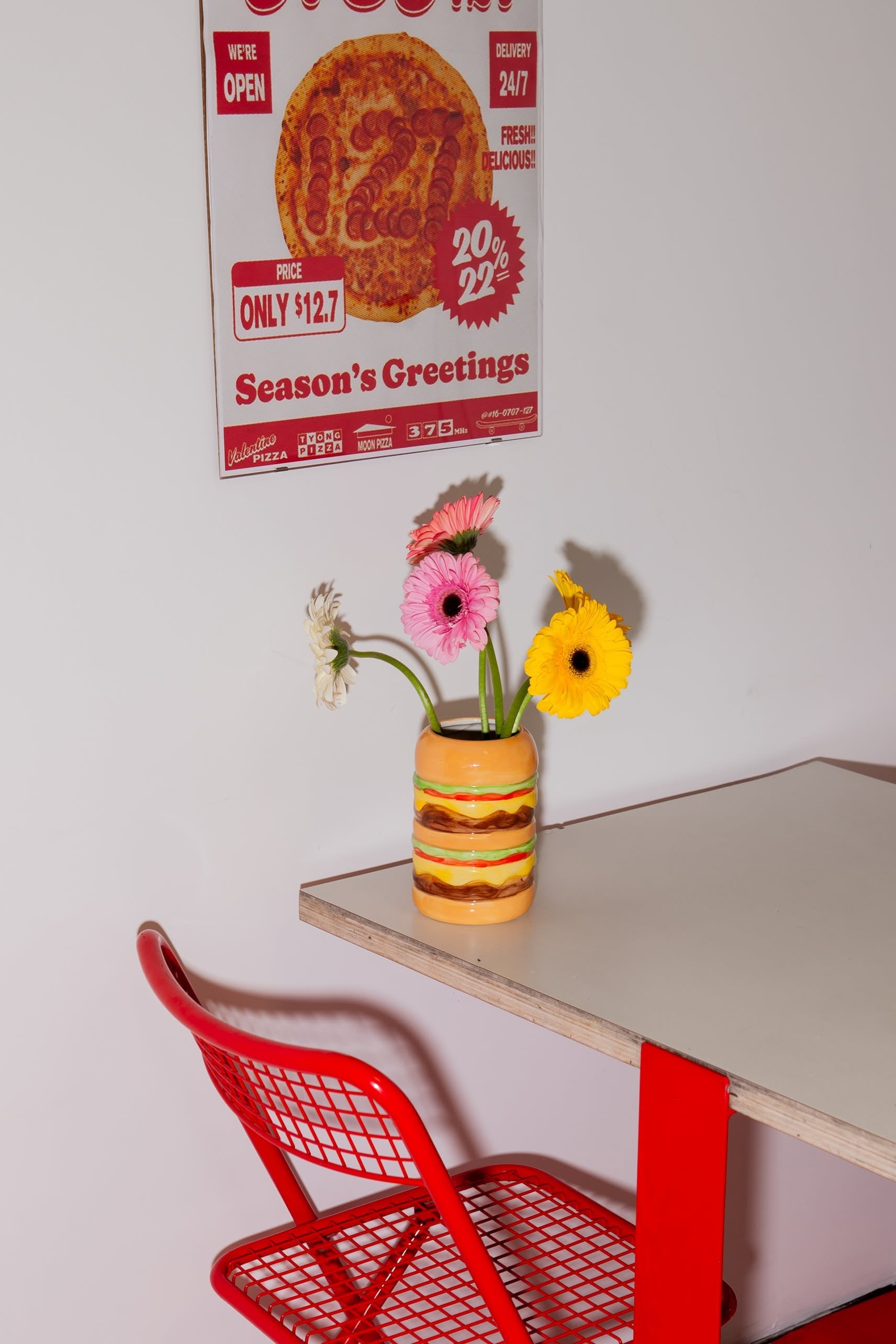 A red metal chair sits by a white table with red legs. On the table is the DOIY Fast Food Burger Vase by DOIY, filled with pink, yellow, and white flowers. A vintage pizza poster on the wall completes the retro-inspired look.