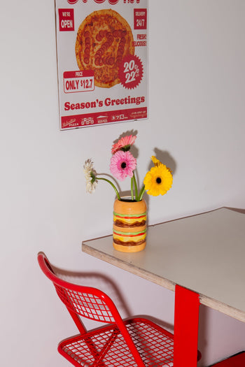 A red metal chair sits by a white table with red legs. On the table is the DOIY Fast Food Burger Vase by DOIY, filled with pink, yellow, and white flowers. A vintage pizza poster on the wall completes the retro-inspired look.