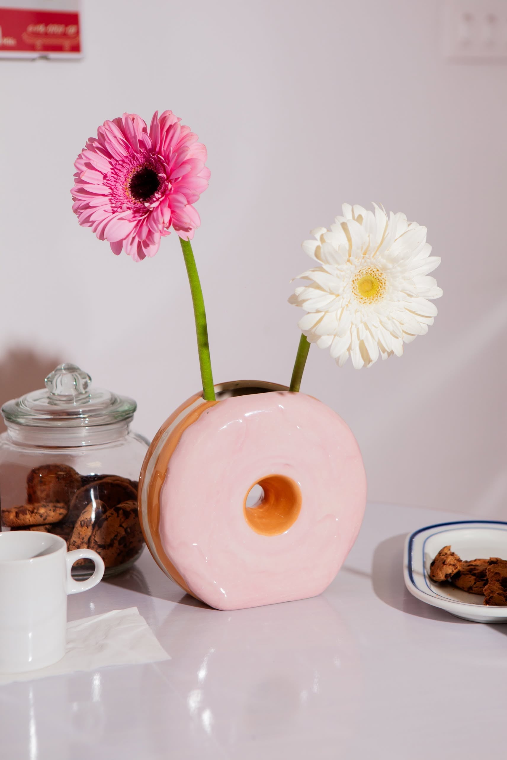 A DOIY Fast Food Donut Vase by DOIY with a glossy pink finish holds a pink and white flower. On the white table are a glass jar of cookies, a white mug, a plate with a cookie, and a napkin.