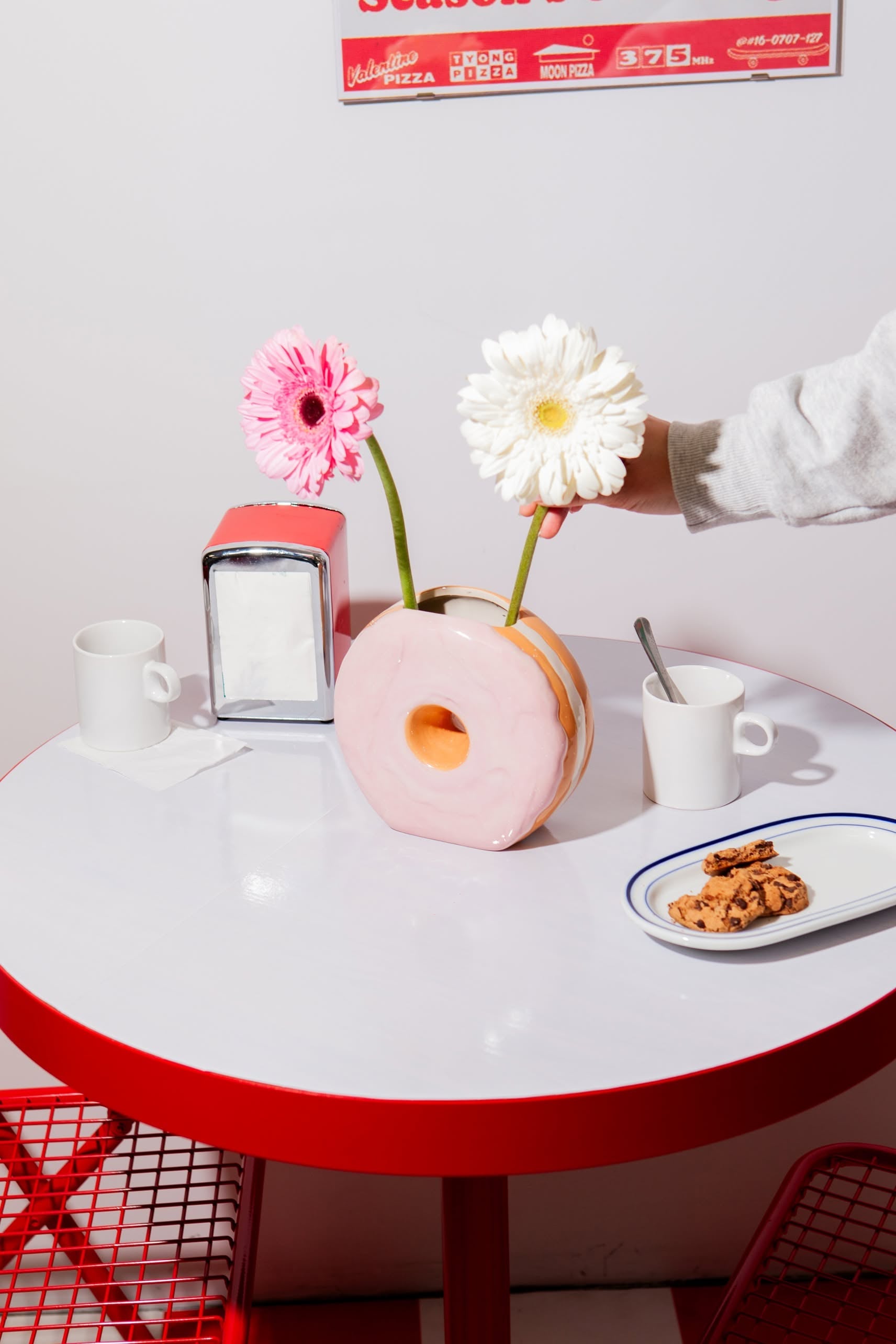 A round table with a red edge and matching chairs holds the DOIY Fast Food Donut Vase by DOIY, displaying two flowers, a napkin holder, two white mugs, and a plate of cookies. A hand adjusts the ceramic vase.