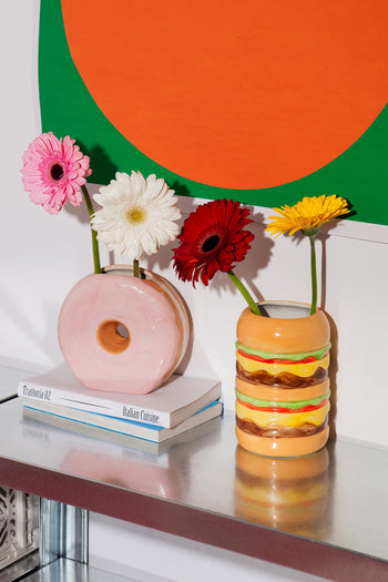 Two quirky ceramic vases—a DOIY Fast Food Donut Vase with a glossy pink glaze and a cheeseburger—hold colorful gerbera daisies on a metallic shelf, flanked by stacked books and an abstract orange and green artwork in the background.