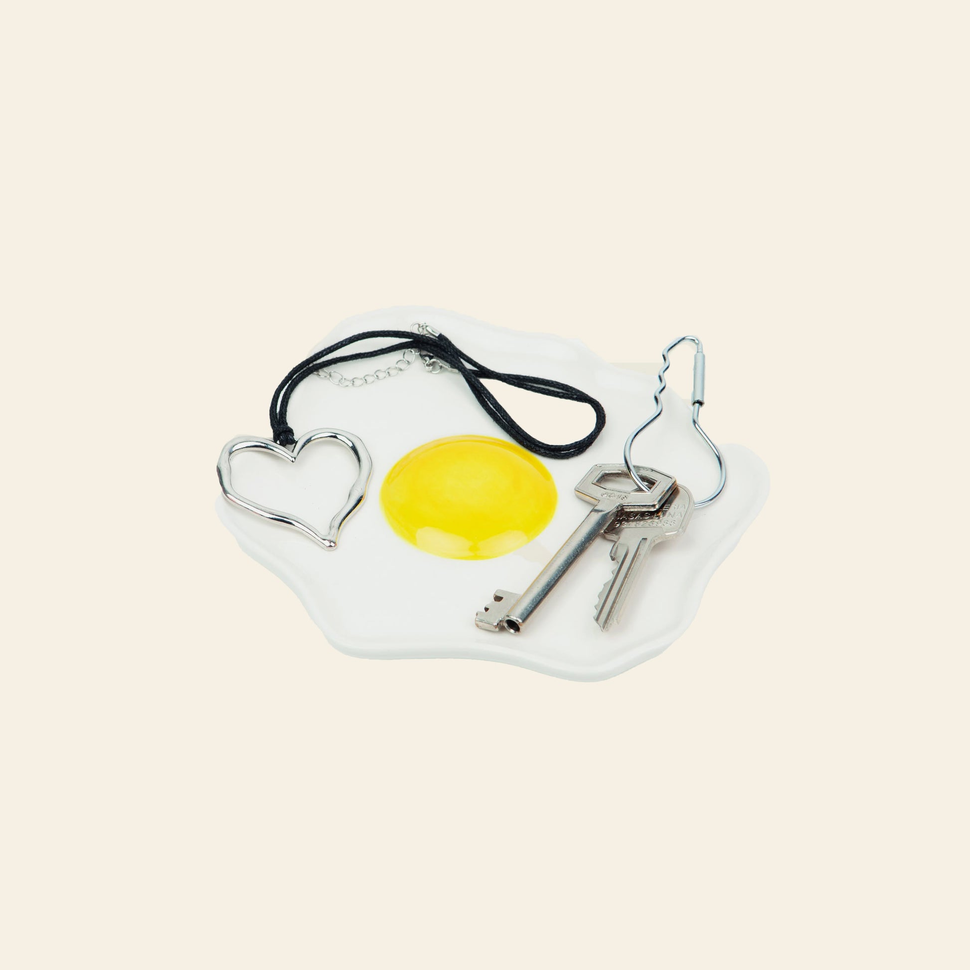 The DOIY Fast Food Fried Egg Trinket Tray by DOIY, with a retro white ceramic design, holds a metal heart keyring, black cord necklace, and two silver keys on a light beige backdrop.