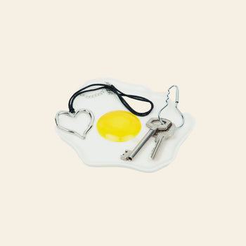The DOIY Fast Food Fried Egg Trinket Tray by DOIY, with a retro white ceramic design, holds a metal heart keyring, black cord necklace, and two silver keys on a light beige backdrop.