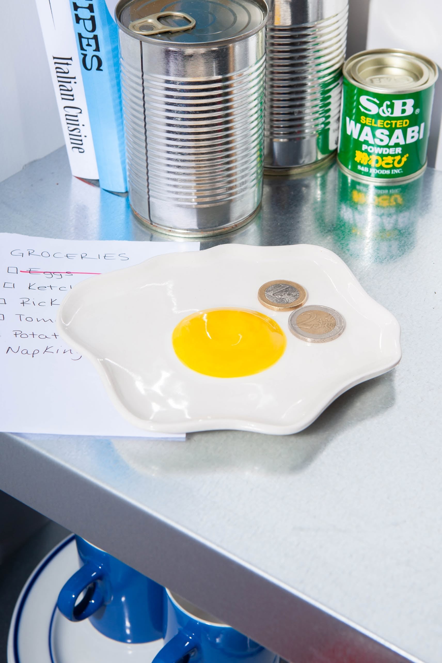 The DOIY Fast Food Fried Egg Trinket Tray by DOIY holds two coins. Behind it are a grocery list, canned goods, a wasabi powder tin, and books on a metallic countertop. Blue cups sit tucked away on the lower shelf.