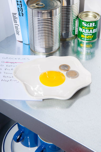 The DOIY Fast Food Fried Egg Trinket Tray by DOIY holds two coins. Behind it are a grocery list, canned goods, a wasabi powder tin, and books on a metallic countertop. Blue cups sit tucked away on the lower shelf.