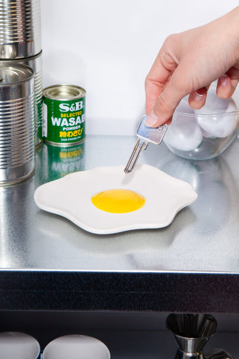 A hand holds a key over the DOIY Fast Food Fried Egg Trinket Tray by DOIY, set on a metallic counter with canned goods, a wasabi tin, and a bowl of eggs—ideal for lovers of vintage-inspired decor.